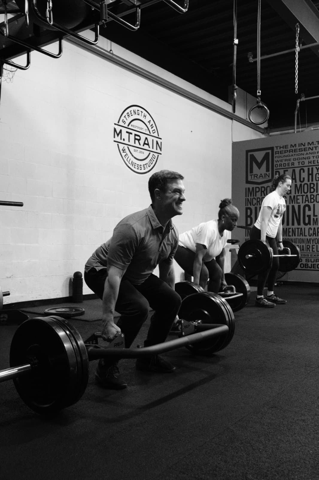 A group class deadlifting together in front of the M.Train logo wall
